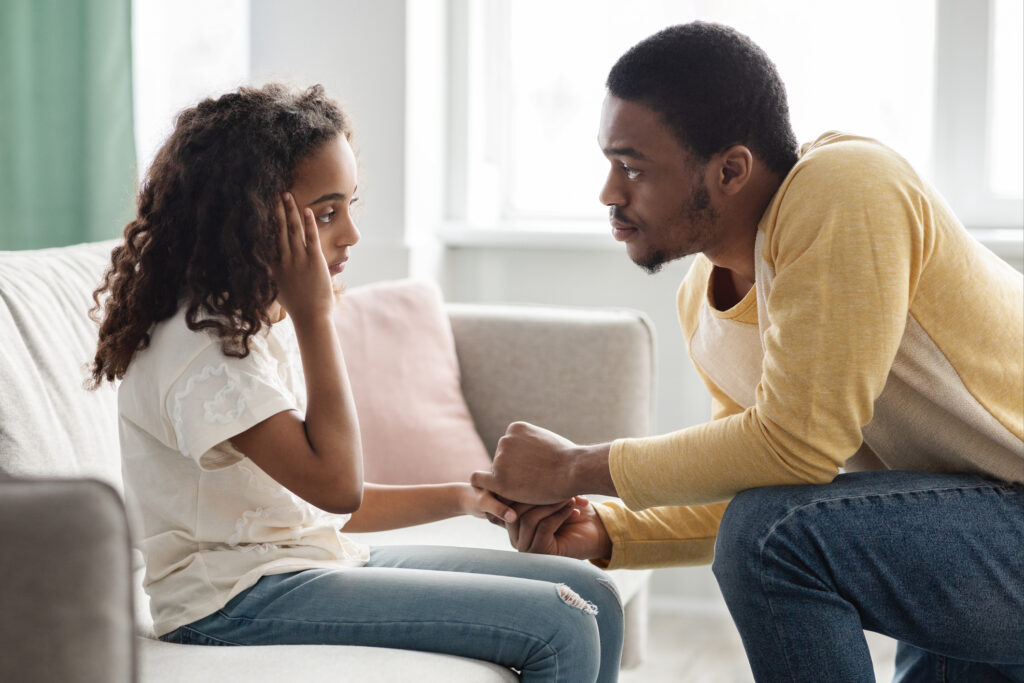 Black father comforting his upset girl kid, side view, home interior. Attentive african american dad consoling sad daughter after quarrel. Fatherhood, parenthood, family relations concept