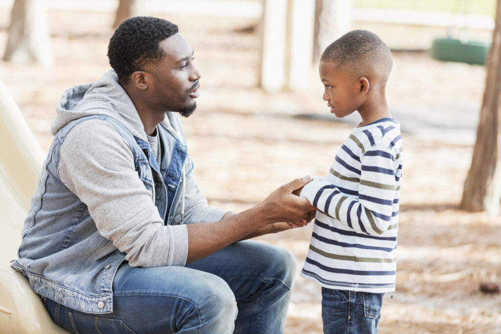 An African-American man in his 30s with a serious expression on his face, talking to his 7 year old son on a playground. They are face to face, and he is holding his hands. The boy may have been naughty and is being disciplined.
